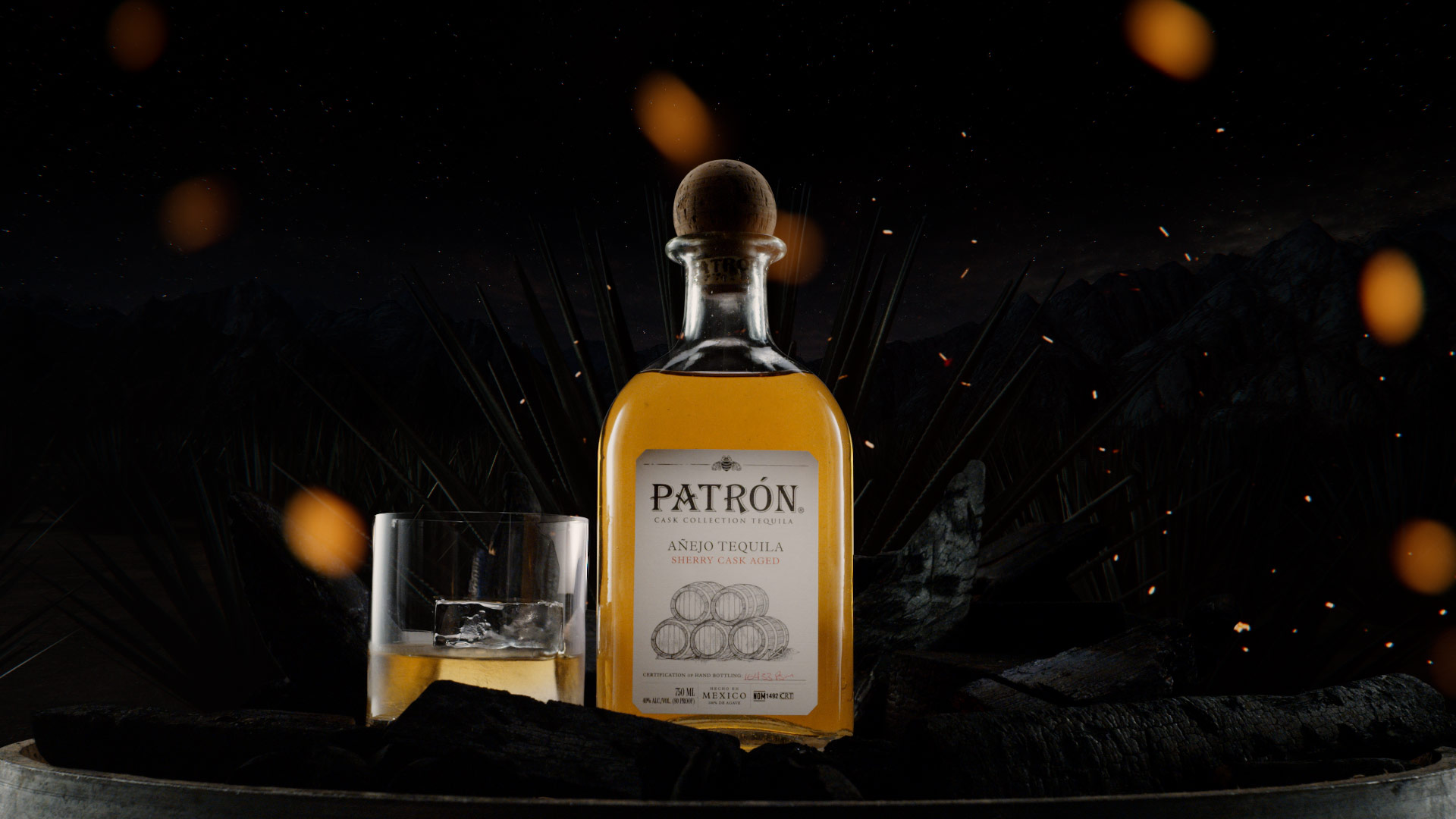A Patron bottle sits on a barel with a glass of tequila next to it. Embers float past the camera.