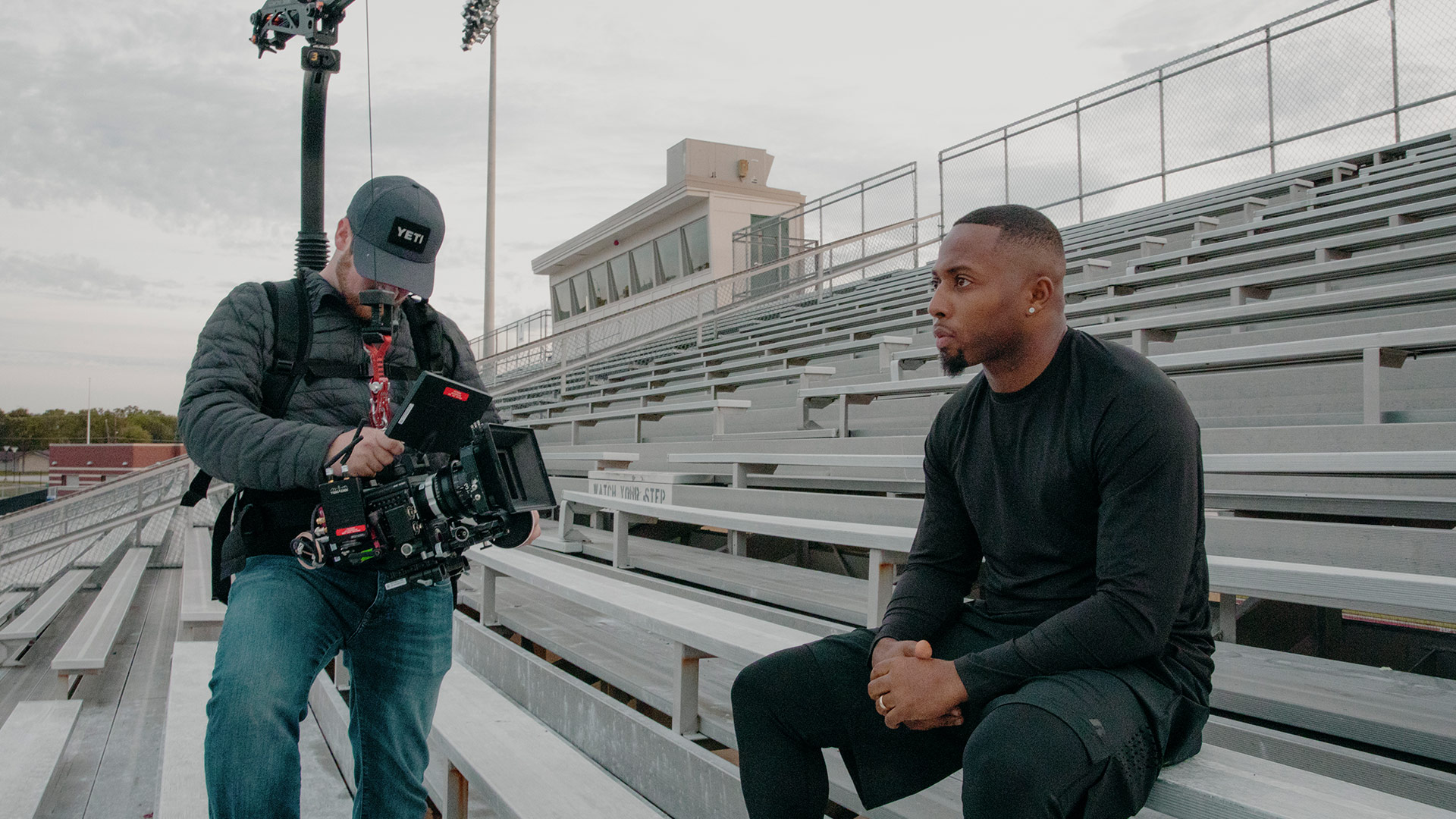 A behind-the-scenes photo of a cinematographer filming a man sitting on bleachers.