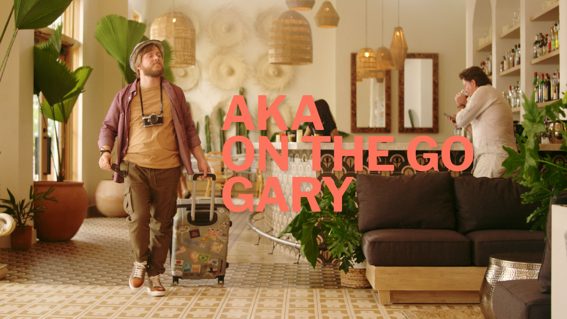 A man with a travel suitcase wlaking through the lobby of a hotel with the text "AKA on the go Gary."